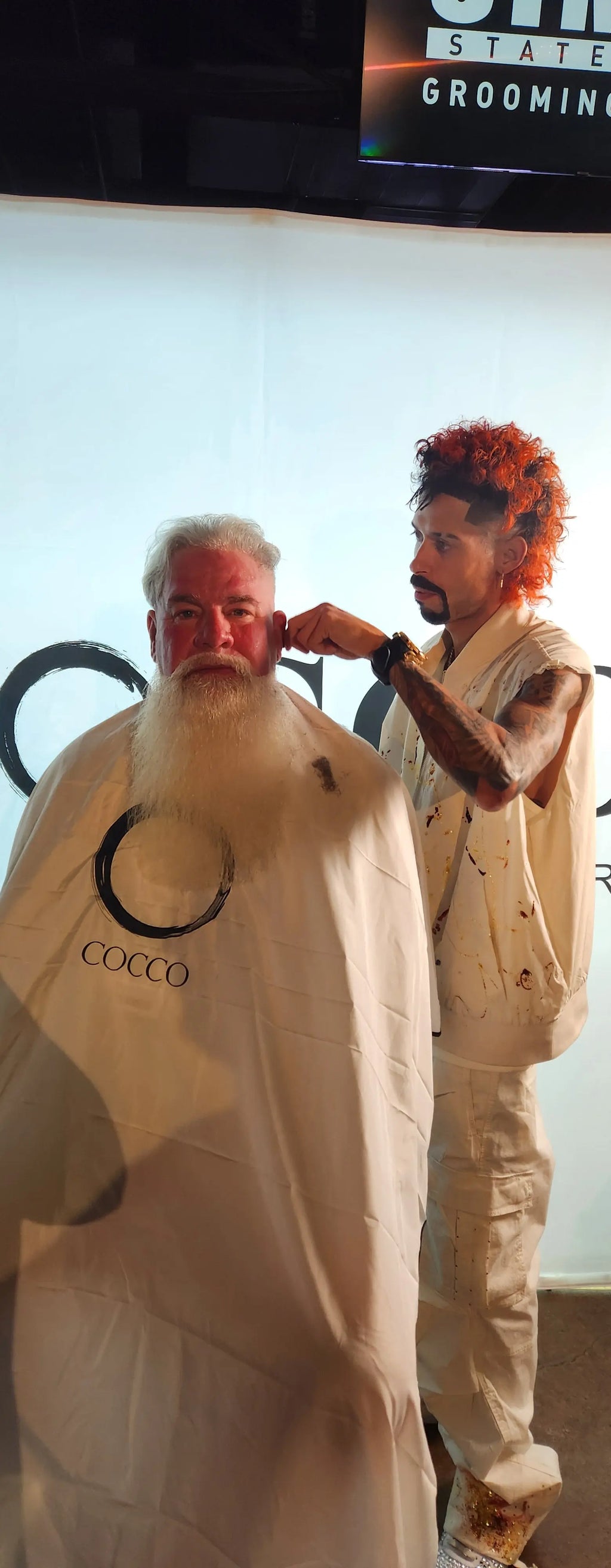 Two people with red hair standing in front of a mirror, one wearing a beige suit and the other in a white outfit.