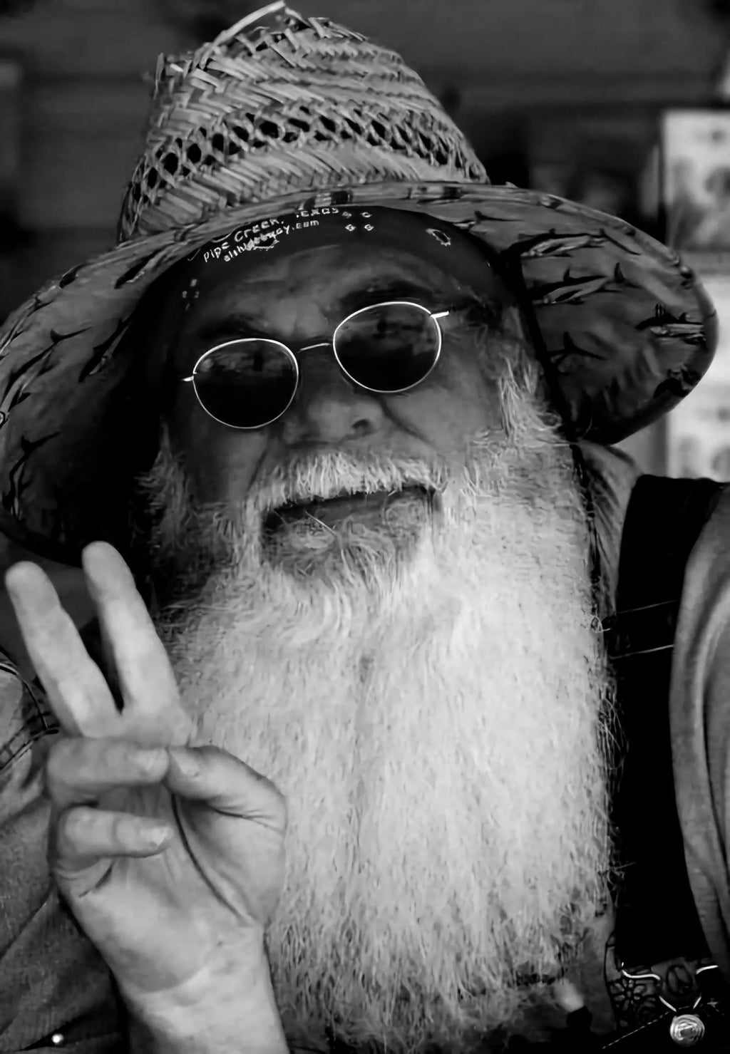 Man with a long beard and sunglasses giving a peace sign, wearing a wide-brimmed hat.