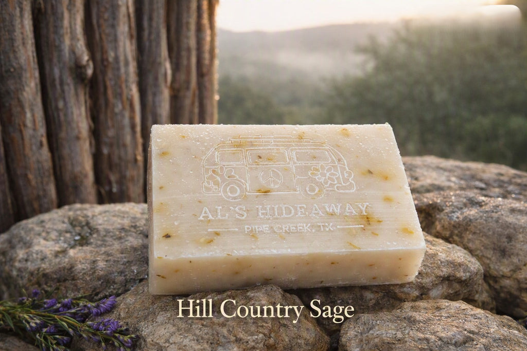 A rectangular bar of soap labeled 'AL'S HIDEAWAY' and 'Hill Country Sage' sits on rocks outdoors, with a blurred natural background and a tree trunk behind it.