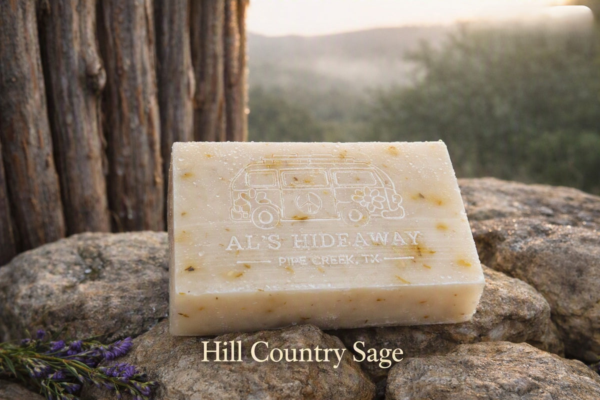 A rectangular bar of soap labeled 'AL'S HIDEAWAY' and 'Hill Country Sage' sits on rocks outdoors, with a blurred natural background and a tree trunk behind it.