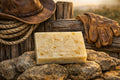 A rectangular bar of soap labeled 'A'L'S HIDEAWAY PIPE CREEK, TX' sits on rocks in front of a rustic wooden fence, with a cowboy hat, rope, and leather gloves styled around it.