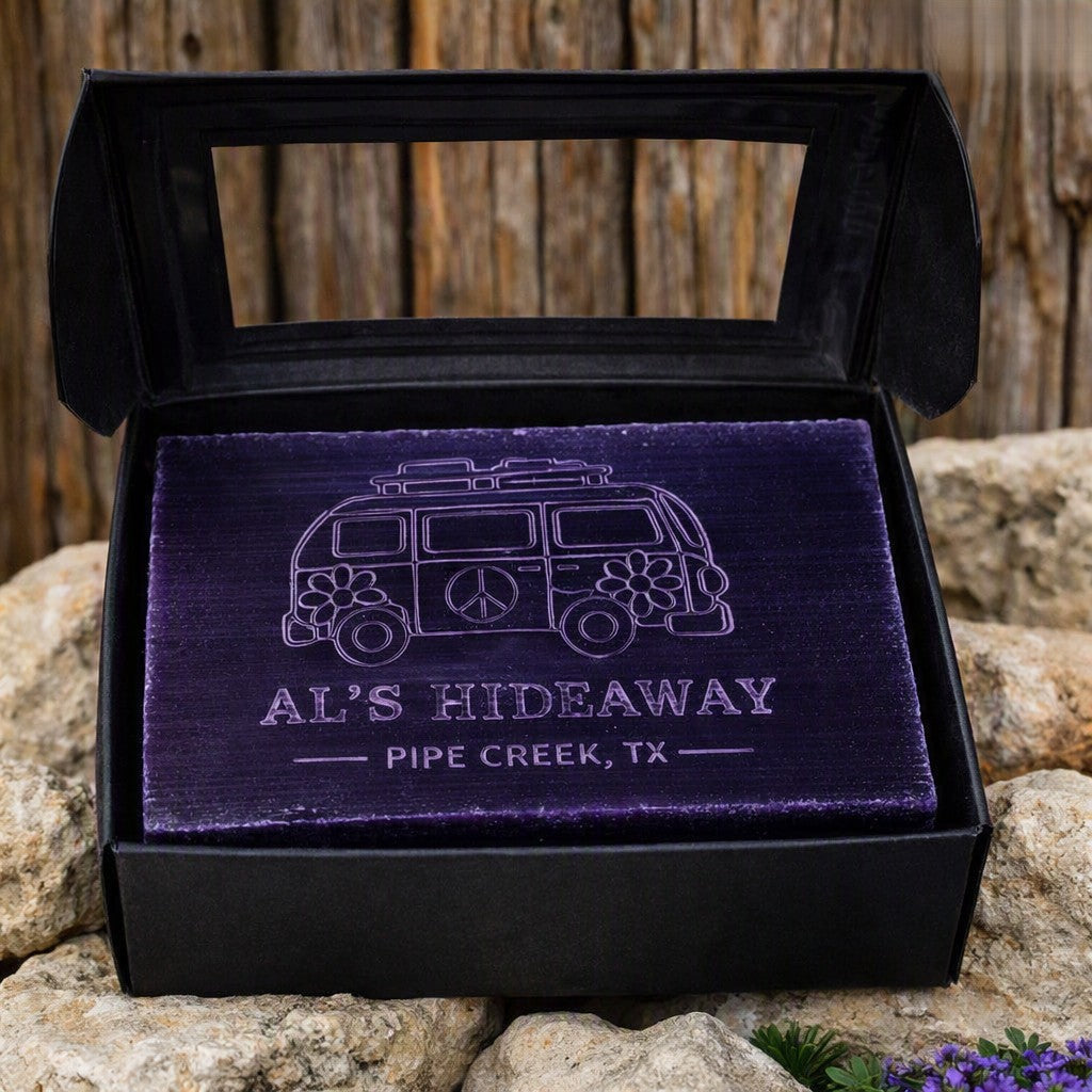 A purple rectangular plaque or gift insert displayed inside an open black box, featuring an engraved retro van illustration and the text 'AL'S HIDEAWAY PIPE CREEK, TX.' The box is set on rocks with a wooden fence background.