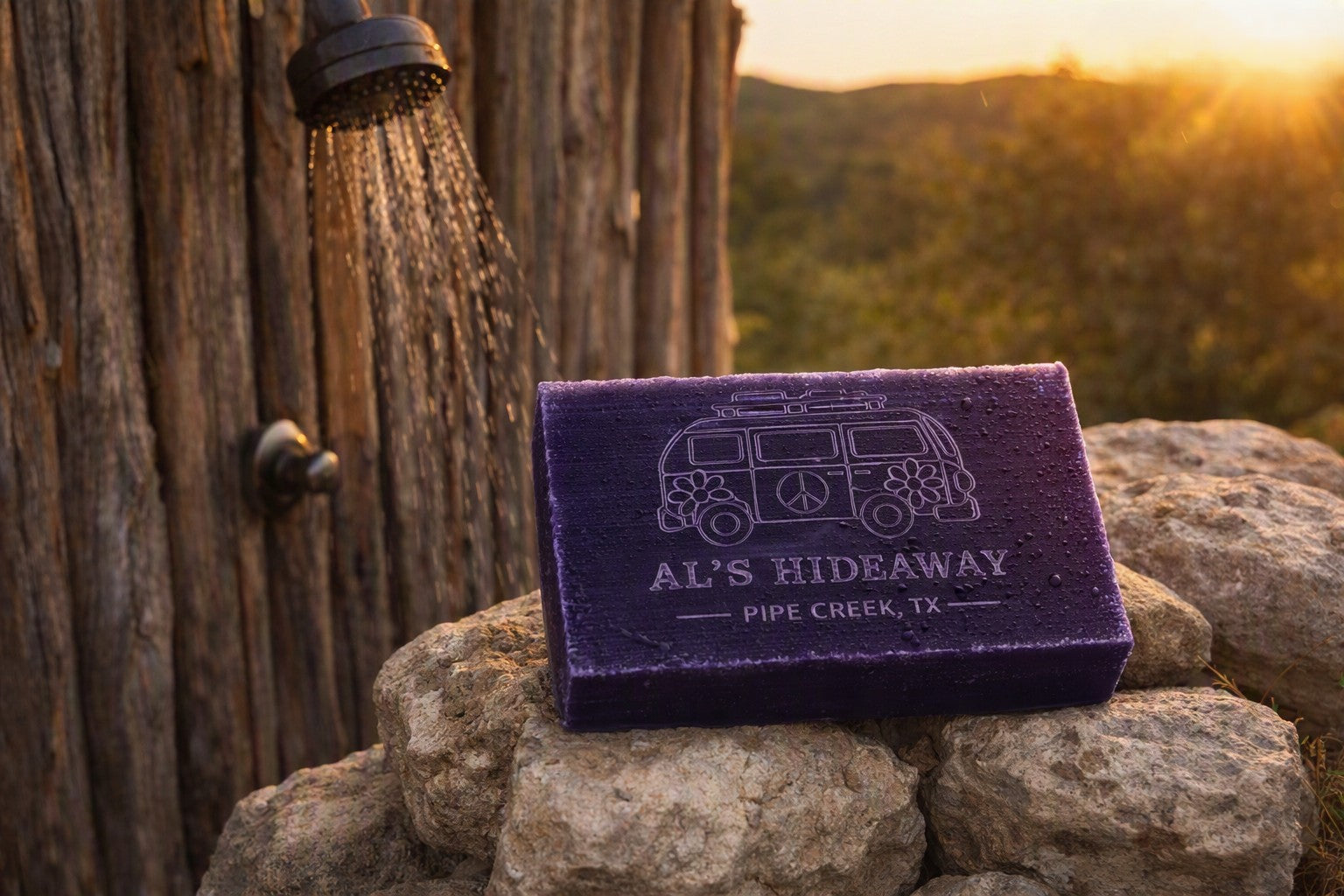 A rectangular purple soap bar engraved with a white van and the text 'AL'S HIDEAWAY PIPE CREEK, TX' sits on rocks beside a rustic wooden wall with a showerhead spraying water, against a warm sunset background.