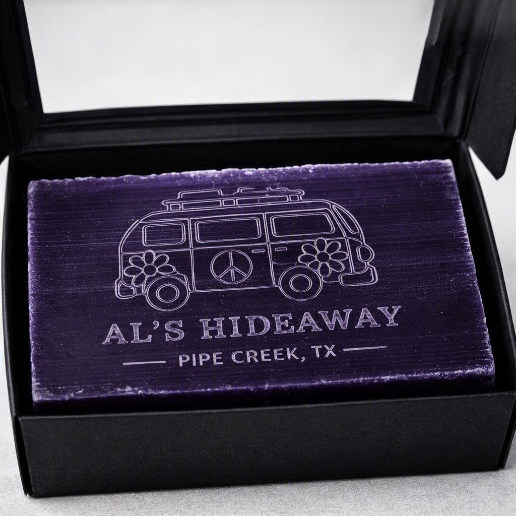 A rectangular purple bar of soap or candle resting in an open black gift box. The soap has a white engraved design of a retro van with flowers and peace signs, along with the text 'AL'S HIDEAWAY' and 'PIPE CREEK, TX'.
