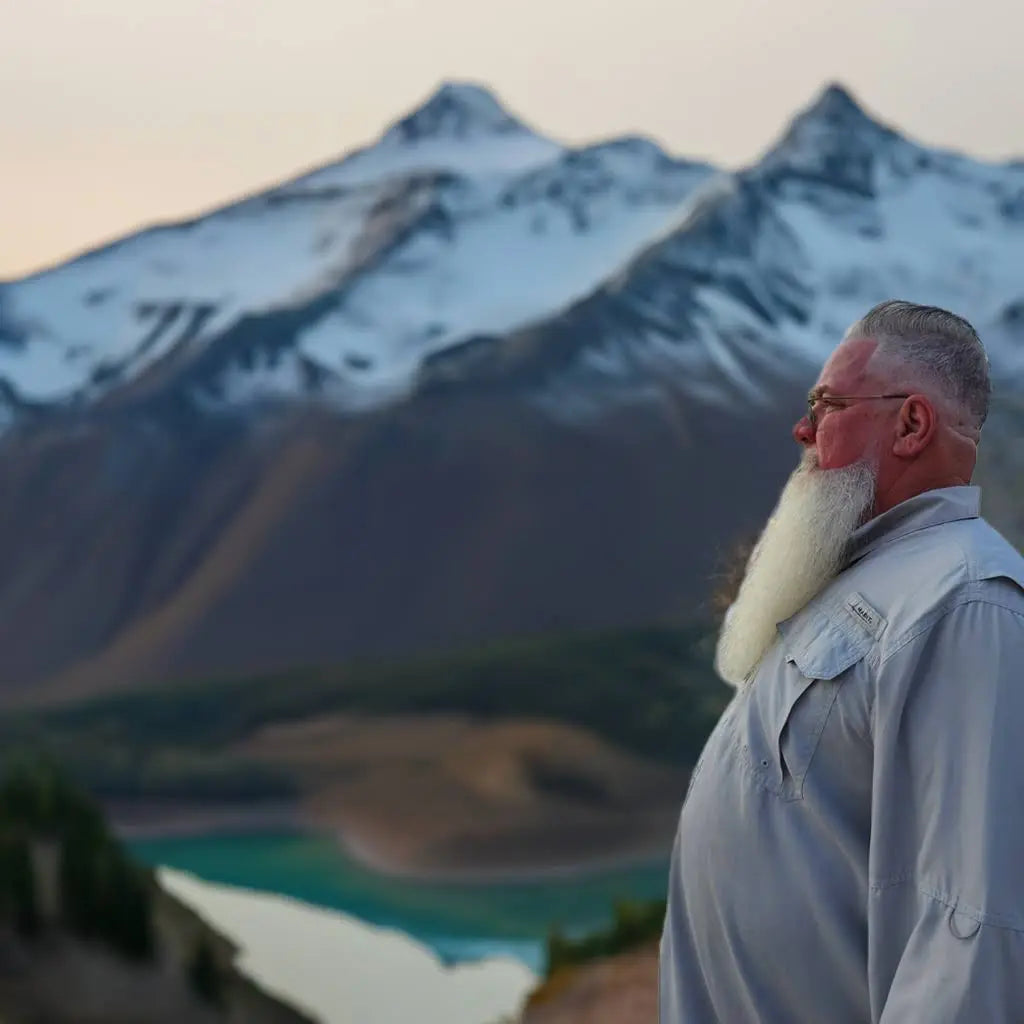 Man with a beard standing in front of a mountain range and lake